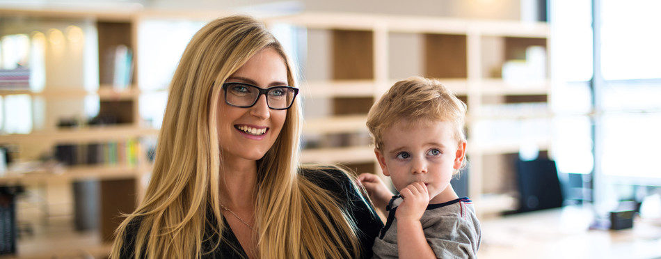 Reconciliation of family and work life: Attractive blond woman in business attire proudly carrying a small boy in her arm in office environment