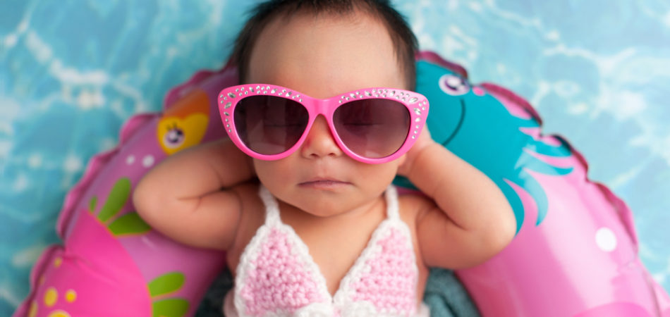 Nine day old newborn baby girl wearing pink sunglasses and a pink and white bikini. She is sleeping on a tiny inflatable swim ring.