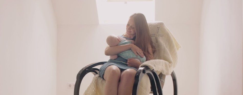 Young beautiful mother smiling at her precious newborn son and lulling him in a rocking chair in a white room in the attic. 4k