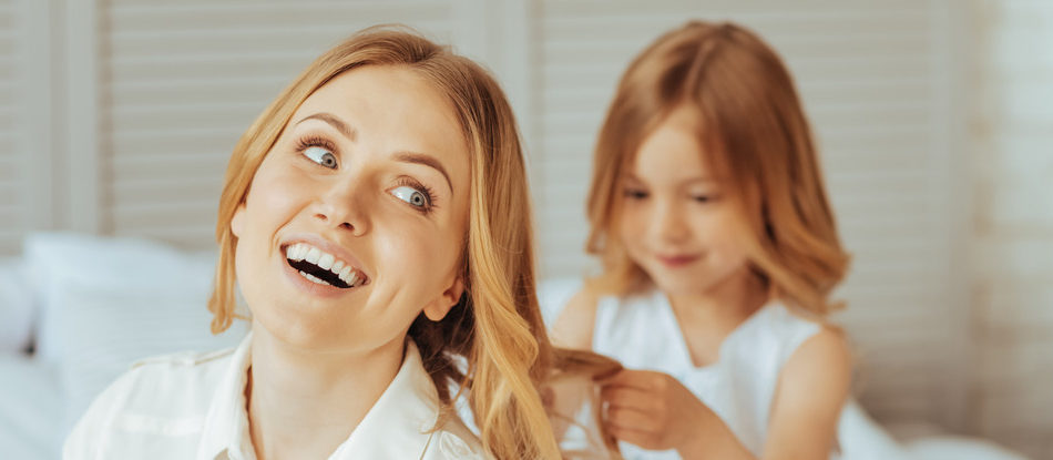 New hairstyle. Delighted positive creative girl sitting behind her mother and holding her hair while doing a new hairstyle for her