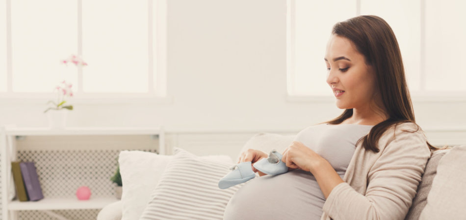 Pregnant woman holding tiny shoes near belly. Smiling expectant lady preparing for baby birth, joying maternity, copy space